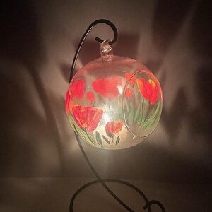 Op de afbeelding: Een glazen bol met een zwarte metalen standaard. De bol is beschilderd met rode en gele bloemen en groene bladeren. De bol wordt van binnenuit verlicht, waardoor een warme gloed ontstaat.