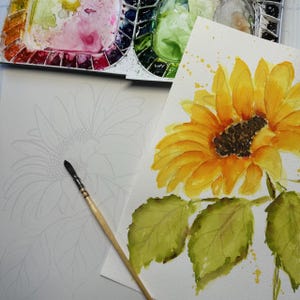 Op de afbeelding: Een aquarel schilderij van een zonnebloem met gele bloemblaadjes en een donkerbruin centrum, naast een gedetailleerde schets van een zonnebloem. Een penseel en een aquarelpalet met diverse kleuren zijn ook zichtbaar.