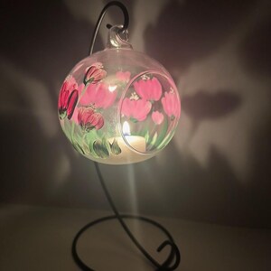 Op de afbeelding: Een heldere glazen bol met een geschilderd ontwerp van roze bloemen en groene bladeren. De bol hangt aan een zwarte metalen standaard en heeft een kleine kaars erin.