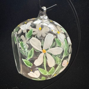 Op de afbeelding: Een heldere glazen bal ornament met handgeschilderde witte bloemen en groene bladeren. De bloemen hebben gele centra.