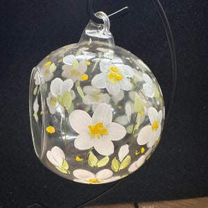 Op de afbeelding: Een helder glazen terrarium met een ronde vorm en een platte bodem. Het terrarium is versierd met witte bloemen met gele centra en groene bladeren. Het heeft een kleine opening en een metalen ophanglus.
