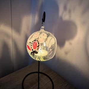 Op de afbeelding: Een heldere glazen bol met een zwarte metalen standaard. De bol heeft een geschilderd vlinderontwerp met rode, witte en zwarte accenten. De bol wordt van binnenuit verlicht, waardoor een warme gloed ontstaat.