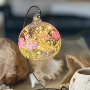 Op de afbeelding: Een heldere glazen bol met roze en groene bloemmotieven, verlicht door warmwitte lichtslingers. De bol hangt aan een zwarte metalen standaard. Decoratieve woonaccessoires.
