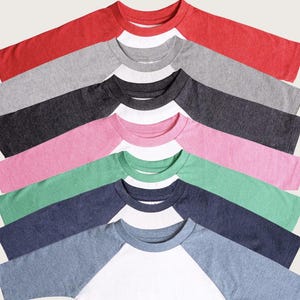May include: A collection of raglan baseball shirts in various colors, including red, gray, black, pink, green, and blue. Each shirt has a white body and colored sleeves. The shirts are arranged in a diagonal pattern, showcasing the different color combinations.