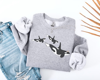 May include: A light gray sweatshirt featuring a graphic of three orcas in black and white. The sweatshirt has a crew neck and long sleeves with white cuffs. A pair of blue jeans with distressed details are visible.