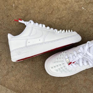 Puede incluir: Zapatillas de cuero blancas con detalles rojos y tachuelas. Las zapatillas tienen cordones blancos con puntas rojas y una suela roja. La palabra "AIR" es visible en el lateral.