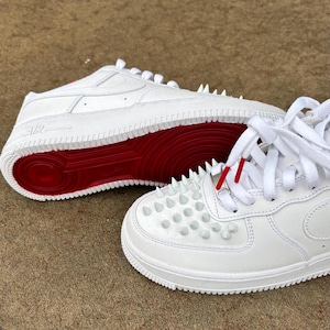 Puede incluir: Zapatillas blancas con suelas rojas y tachuelas plateadas en la puntera. Los zapatos tienen cordones blancos y puntas de cordones rojas. Las suelas son de color rojo brillante y los zapatos son de material blanco liso.