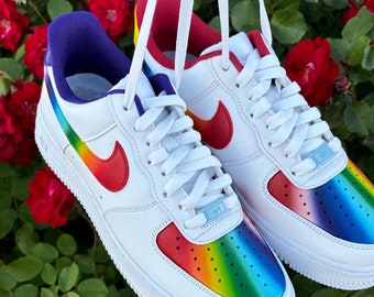May include: A pair of white sneakers featuring a vibrant rainbow gradient on the perforated toe box and side panels. The shoes have red swoosh logos, white laces, and light blue lace dubraes labeled AFI. The inner collars are purple and red.