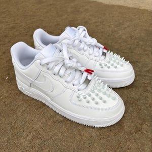 Puede incluir: Un par de zapatillas de cuero blanco con cordones blancos y un dise&ntilde;o de puntera tachonada. Las zapatillas presentan el logotipo de Nike swoosh en el lateral y detalles rojos en los cordones.