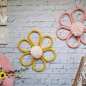 May include: Two macrame flower wall hangings, one in pink and one in yellow. The pink flower has a white pom-pom centre, while the yellow flower has a white fluffy centre.