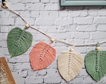 May include: A macrame garland with five green, pink, and cream leaves hanging on a white brick wall. Each leaf is connected by a white string with a wooden bead in between.