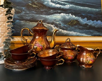 May include: A tea set featuring a deep burgundy and gold luster finish with ornate gold filigree detailing. The set includes a teapot with a lid, a lidded sugar bowl, a creamer, and two teacups with matching saucers, all decorated with colorful pastoral scenes.