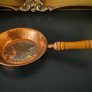 May include: A round copper pan featuring a hammered texture on the interior surface and a long, cylindrical wooden handle with three decorative grooves and a small circular hole at the end for hanging purposes.