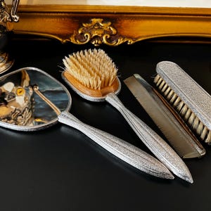 Puede incluir: Un set de tocador que incluye un espejo de mano, un cepillo para el cabello con cerdas claras, un peine de dientes finos y un cepillo para ropa. Cada pieza presenta un mango o respaldo plateado texturizado con un acabado metálico uniforme.