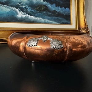 May include: A round copper bowl with a polished metallic finish and a slightly weathered patina. The exterior features a decorative silver-toned metal appliqué depicting a cluster of grapes with two leaves centred on the side of the bowl.