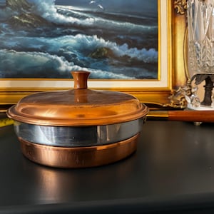 May include: A round copper pan with a matching lid and a long wooden handle. The pan features a polished copper exterior, a silver-coloured metal band around the centre, and a small copper knob on the lid. The handle is brown wood with a brass collar.