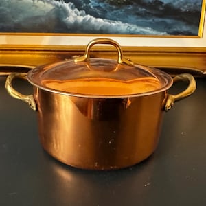 Puede incluir: Una olla de cobre pulido con tapa a juego y dos asas laterales de latón. La olla presenta un cuerpo cilíndrico con una superficie lisa y reflectante, además de un asa curva en la parte superior de la tapa, con un acabado metálico cálido.