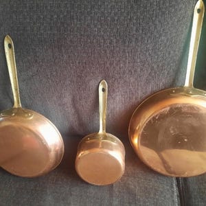 Op de afbeelding: Drie koperen pannen met lange messing handgrepen, elk voorzien van een klein rond gat aan het uiteinde van de steel om op te hangen. De pannen variëren in grootte, met twee koekenpannen met platte bodem en één kleinere, diepere steelpan.