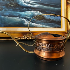 May include: A copper watering can featuring a long, slender spout and a curved handle. The cylindrical body is decorated with a detailed embossed frieze depicting horses and figures in motion. The metal surface has a warm, metallic finish.