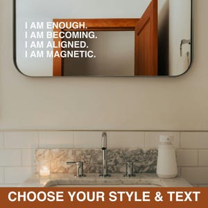May include: A rectangular wall mirror featuring white adhesive text that reads: I AM ENOUGH. I AM BECOMING. I AM ALIGNED. I AM MAGNETIC. The mirror has rounded corners and reflects a wooden door frame, positioned above a marble sink with a faucet.
