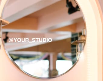 May include: A round mirror with a white frame and a light pink background. The mirror is lit with a string of white lights. The text "@YOUR_STUDIO" is visible in the reflection.