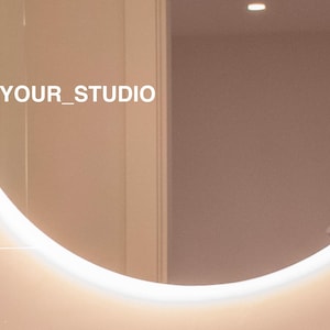 May include: A round, white-rimmed mirror with a light brown background. The mirror reflects a room with a white door and a light fixture. The text "@YOUR_STUDIO" is visible in the reflection.