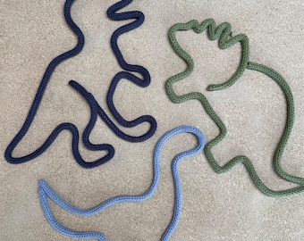 May include: Three dinosaur-shaped decorations made from a thick, textured rope. One is navy blue, one is light blue, and the other is green. The dinosaurs are arranged on a neutral, light brown surface.