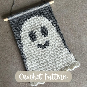May include: A crocheted ghost wall hanging in grey and white. The ghost has black eyes and a smiling mouth. The banner is attached to a wooden dowel and has the words "Crochet Pattern" at the bottom.