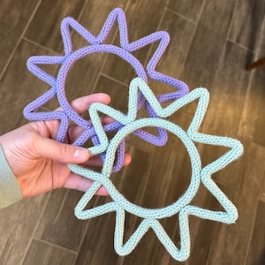 May include: Two crocheted sun shapes, one in light purple and one in light green, are held in a hand. The suns are both circular with points radiating out from the circle.