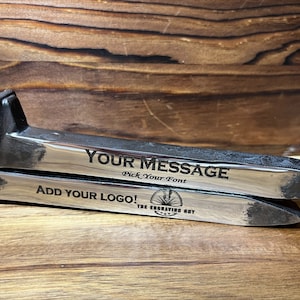 May include: Two metal horseshoe nails with engraved text: "YOUR MESSAGE" and "ADD YOUR LOGO!". The nails are silver with dark gray accents and are displayed on a wooden surface. The text "Pick Your Font" is also visible.