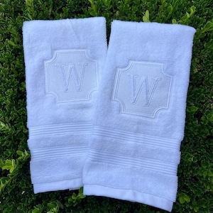 May include: Two white terry cloth hand towels featuring a centered embroidered monogram of the letter W enclosed within a decorative square frame with rounded corners. Each towel has a series of horizontal ribbed bands near the bottom edge.