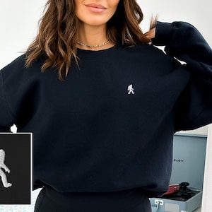 Peut inclure: Sweat-shirt noir avec une image brodée blanche d'un Bigfoot marchant. Le sweat-shirt a un col rond et des manches longues.