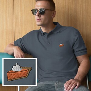 May include: A dark gray polo shirt with a small embroidered image of a pumpkin pie slice with whipped cream. The shirt has a classic collar and button closure. The person is wearing sunglasses.