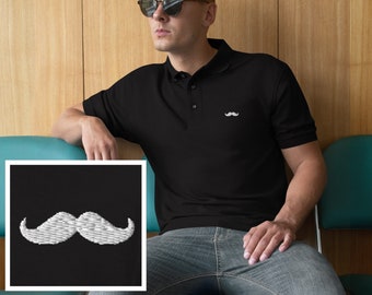 May include: A black polo shirt with a white embroidered mustache on the left chest. The shirt is worn by a person wearing sunglasses and sitting on a teal chair.