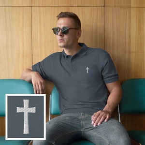 May include: A dark blue polo shirt with a white embroidered cross on the left chest. The shirt is worn by a person wearing sunglasses and jeans.