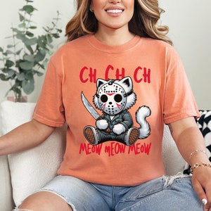 May include: A peach-colored t-shirt with a graphic of a white cat wearing a hockey mask and holding a knife. The text "CH CH CH MEOW MEOW MEOW" is printed below the cat.