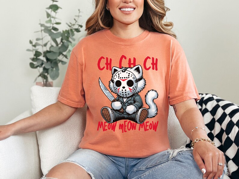May include: A peach-colored t-shirt with a graphic of a white cat wearing a hockey mask and holding a knife. The text "CH CH CH MEOW MEOW MEOW" is printed below the cat.