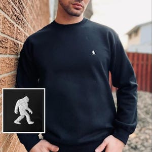 Peut inclure: Un sweat-shirt noir à manches longues et col rond, orné d'une petite broderie blanche représentant une silhouette de Bigfoot en marche sur la poitrine gauche. Le vêtement présente un col, des poignets et un ourlet côtelés.