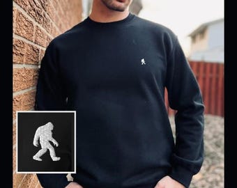 May include: A black long-sleeve crewneck sweatshirt featuring a small, white embroidered graphic of a walking Bigfoot figure on the upper left chest. The sweatshirt has a ribbed neckline, cuffs, and hem, providing a clean and simple design.