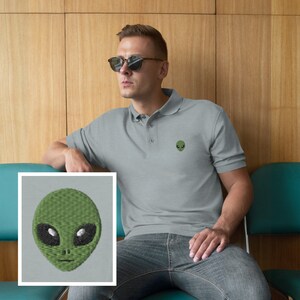 Pu&ograve; includere: Una polo grigia con un viso di alieno verde ricamato sul petto sinistro. L'alieno ha grandi occhi neri e una piccola bocca.