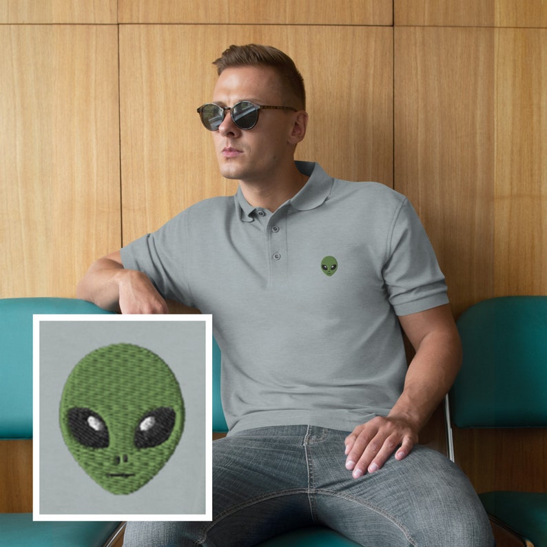 Pu&ograve; includere: Una polo grigia con un viso di alieno verde ricamato sul petto sinistro. L'alieno ha grandi occhi neri e una piccola bocca.