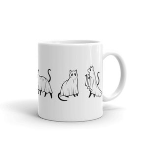 May include: White ceramic coffee mug with a black line drawing of four cats dressed as ghosts. The cats are in various poses, including standing, sitting, and walking.
