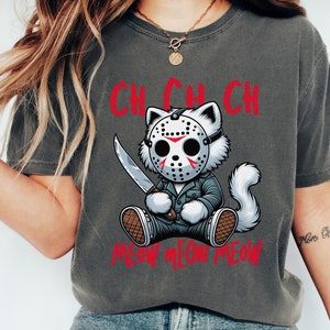 May include: A gray t-shirt with a cartoon cat wearing a hockey mask and holding a knife. The text "Ch Ch Ch Meow Meow Meow" is printed on the shirt.