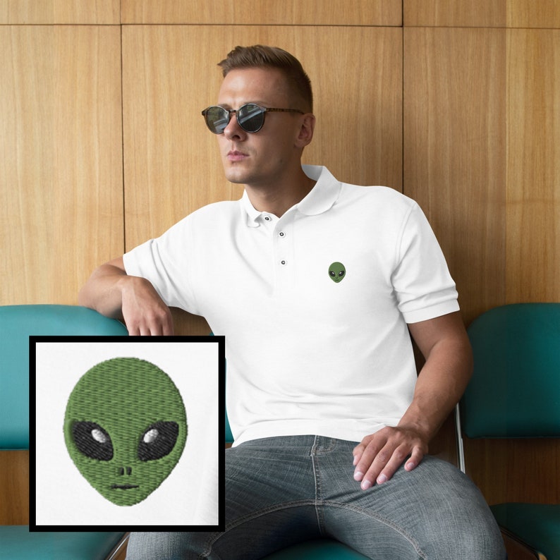 Pu&ograve; includere: Polo bianco con un viso di alieno verde ricamato sul petto sinistro. L'alieno ha grandi occhi neri e una piccola bocca.