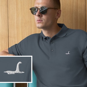 Peut inclure: Un polo bleu foncé avec une image brodée blanche du monstre du Loch Ness. Le polo a un col boutonné et des manches courtes.