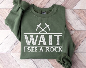 May include: A green sweatshirt with a white graphic that says "WAIT I SEE A ROCK" with two crossed pickaxes above the text.