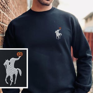 以下が含まれることがあります： カボチャを持った首なし騎士の刺繍デザインが施された黒いクルーネックのスウェットシャツ。デザインは白で、オレンジ色のカボチャが付いています。スウェットシャツは無地です。