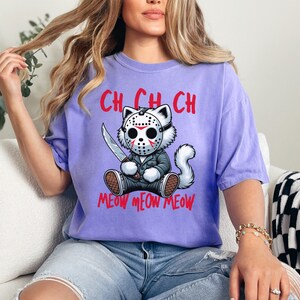 May include: A purple t-shirt with a cartoon cat wearing a hockey mask and holding a knife. The text "Ch Ch Ch Meow Meow Meow" is printed on the shirt.