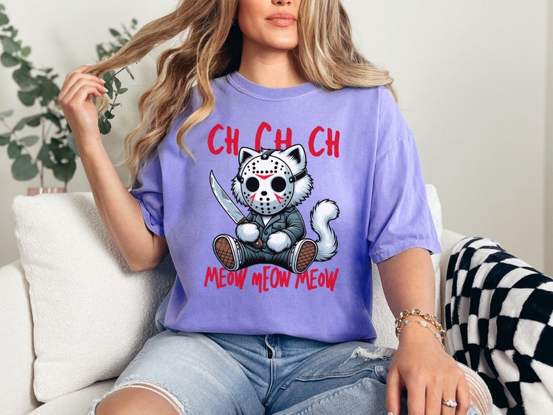 May include: A purple t-shirt with a cartoon cat wearing a hockey mask and holding a knife. The text "Ch Ch Ch Meow Meow Meow" is printed on the shirt.