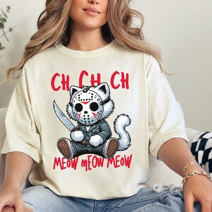 May include: A cream-colored t-shirt with a cartoon cat wearing a hockey mask and holding a knife. The text "Ch Ch Ch Meow Meow Meow" is printed on the shirt.